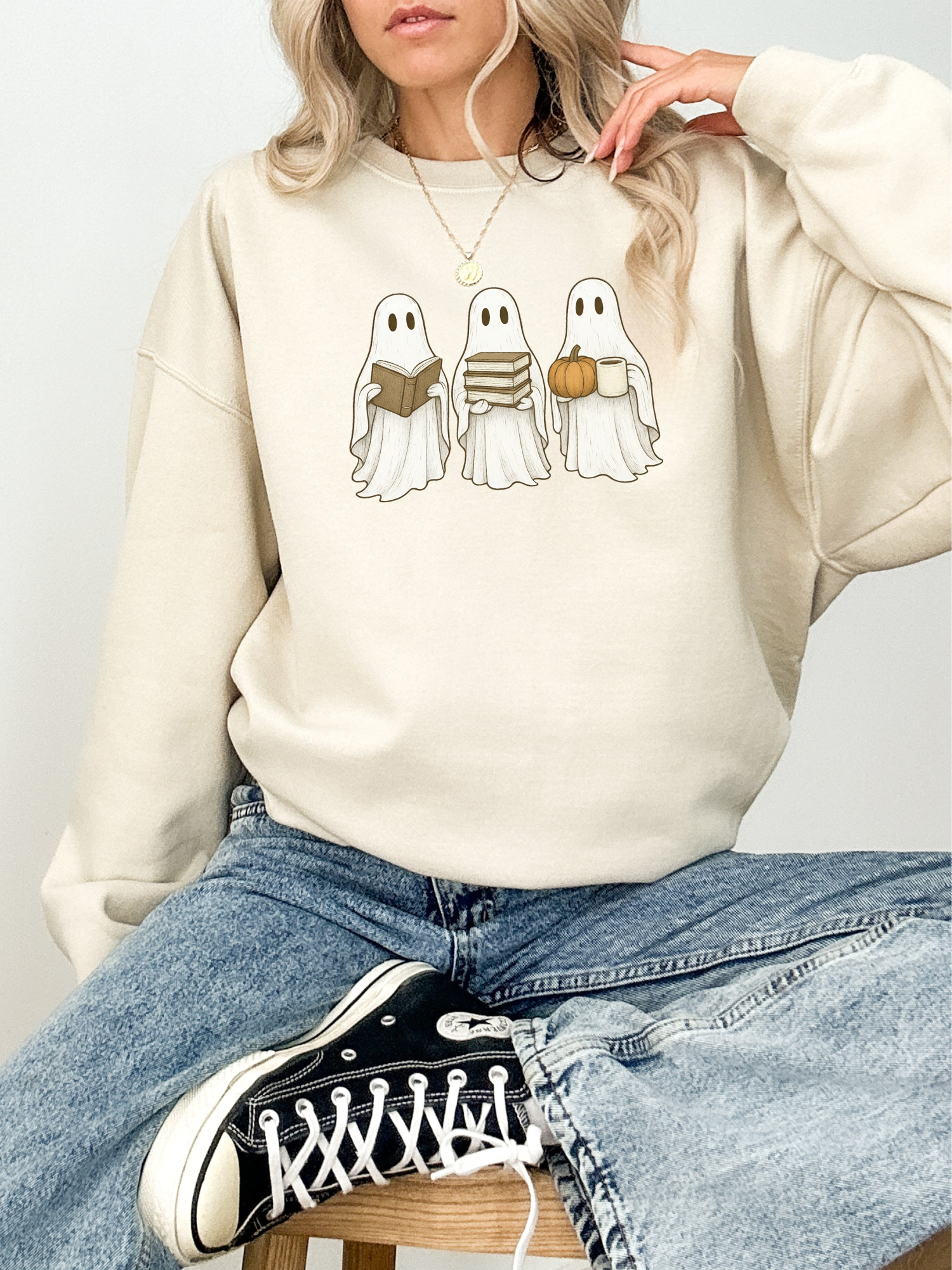 woman in a sand-colored sweatshirt with cartoon ghosts holding fall-themed bookish items, styled for Halloween