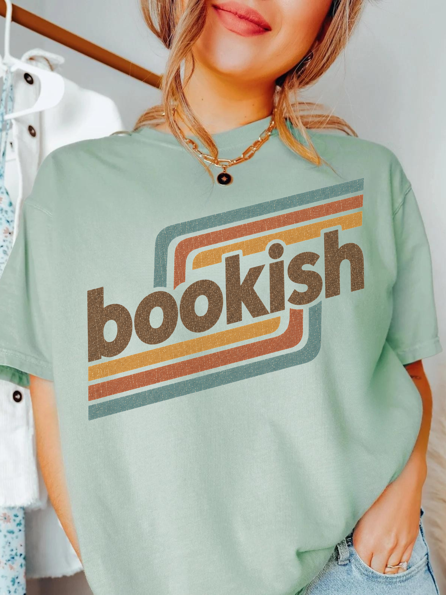 A woman wearing a bay green graphic t-shirt with the word 'bookish' in brown retro lettering over orange and teal stripes. She is wearing a gold necklace and blue jeans, with a clothing rack visible in the background.