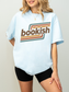 A woman wearing a chambray oversized graphic T-shirt with the word 'bookish' in brown text over retro orange, yellow, and teal stripes, paired with black bike shorts against a light gray studio background.