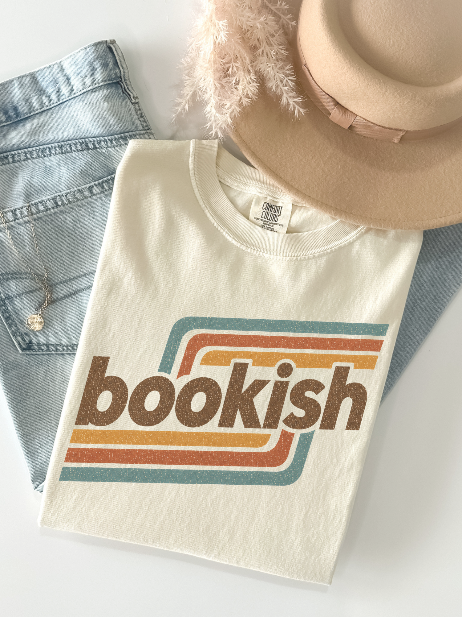 A folded ivory graphic T-shirt with the word 'bookish' in brown over retro orange, yellow, and teal stripes, styled on a white surface with light-wash denim jeans, a beige hat, and soft beige feathers.