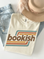 A folded ivory graphic T-shirt with the word 'bookish' in brown over retro orange, yellow, and teal stripes, styled on a white surface with light-wash denim jeans, a beige hat, and soft beige feathers.