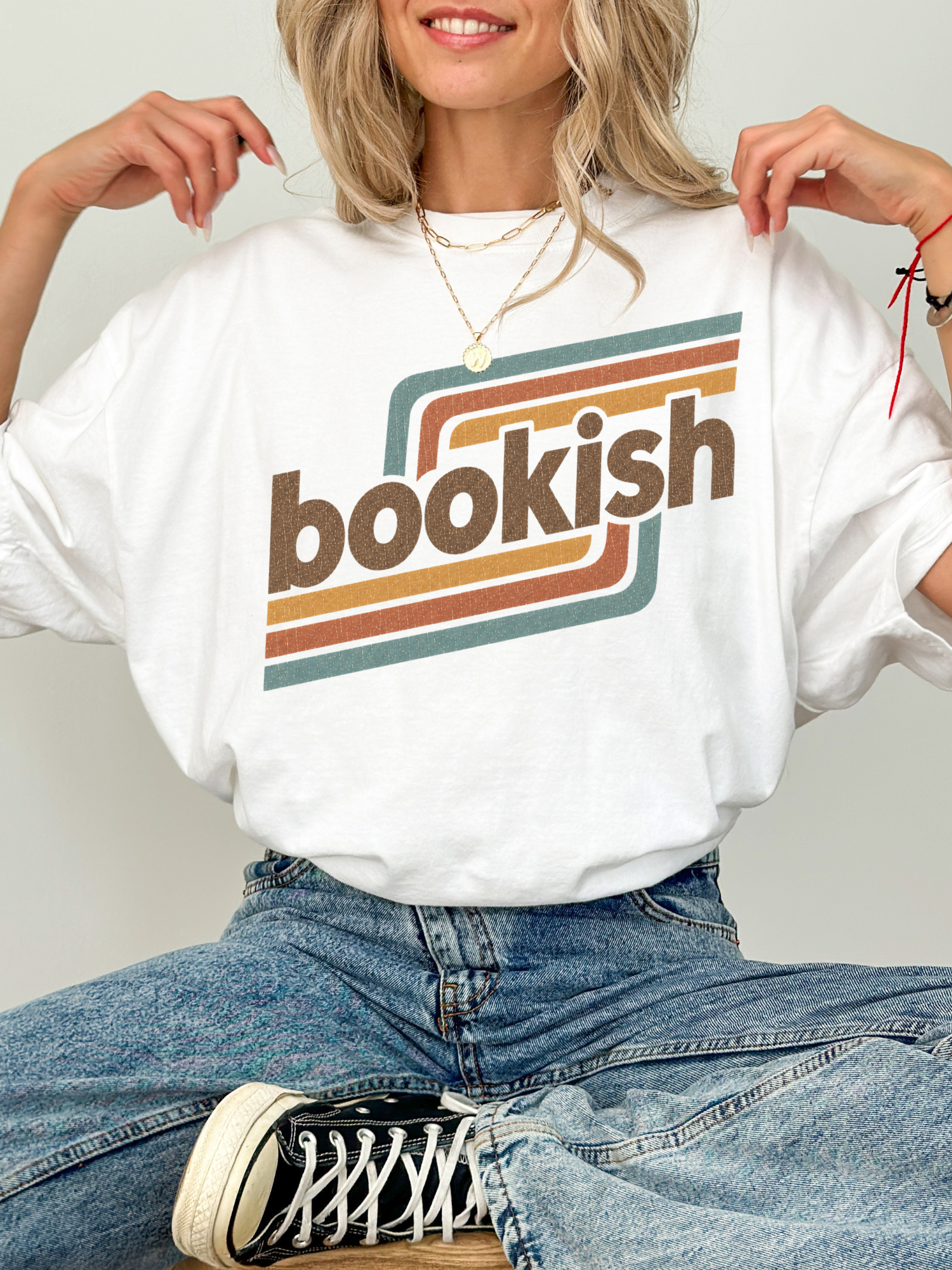 A woman wearing a white oversized graphic T-shirt with the word 'bookish' in brown letters over retro brown, orange, and teal stripes. She is also wearing blue jeans and a black-and-white sneaker, posed against a light gray studio background.