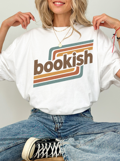 A woman wearing a white oversized graphic T-shirt with the word 'bookish' in brown letters over retro brown, orange, and teal stripes. She is also wearing blue jeans and a black-and-white sneaker, posed against a light gray studio background.