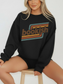 A woman wearing a black crewneck sweatshirt with a retro-style 'bookish' graphic in brown, orange, and teal, sitting on a wooden stool against a plain white background.