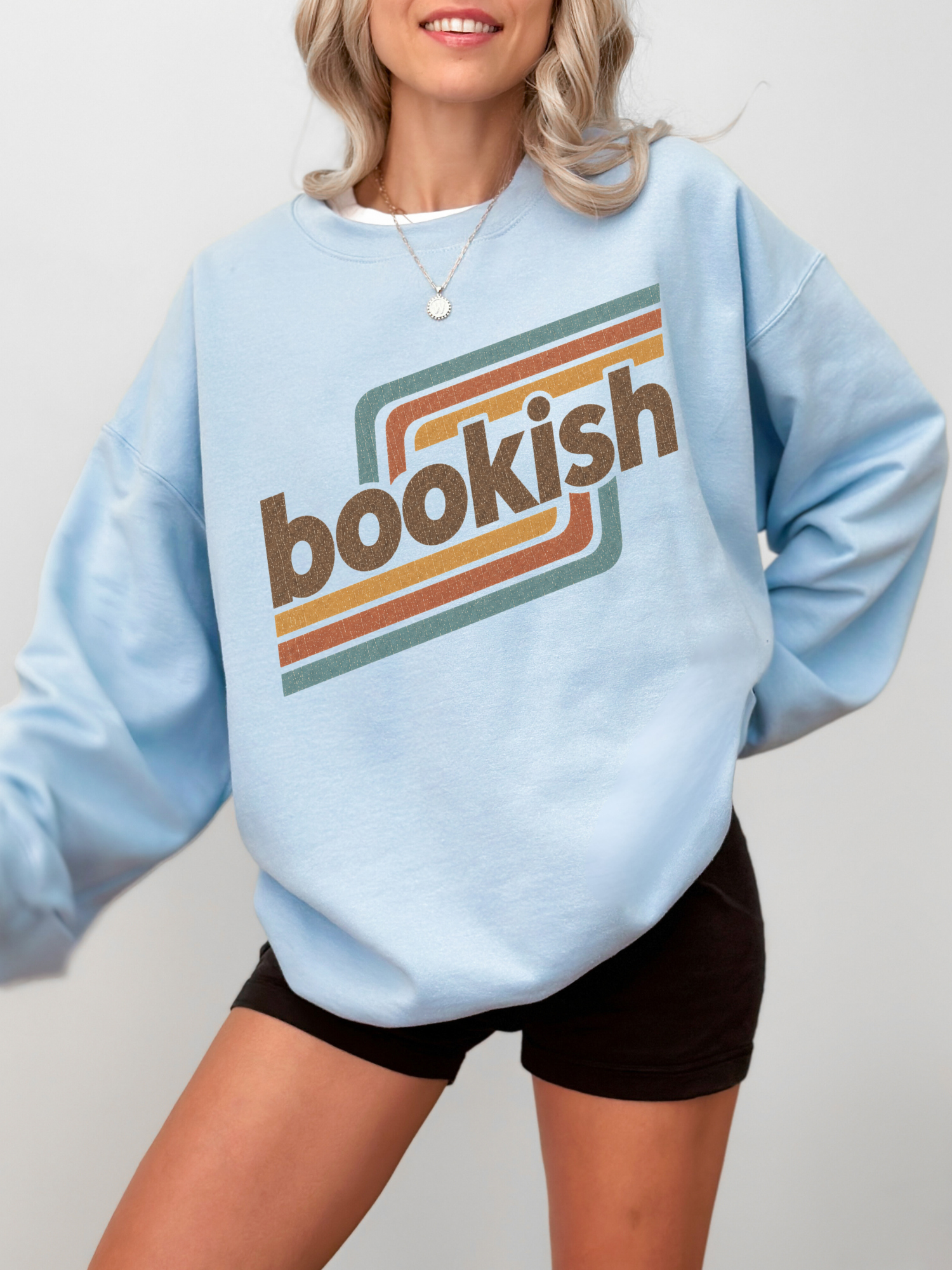 A woman wearing a light blue crewneck sweatshirt with a retro-style 'bookish' graphic in brown, orange, and teal, paired with black shorts against a light gray background.
