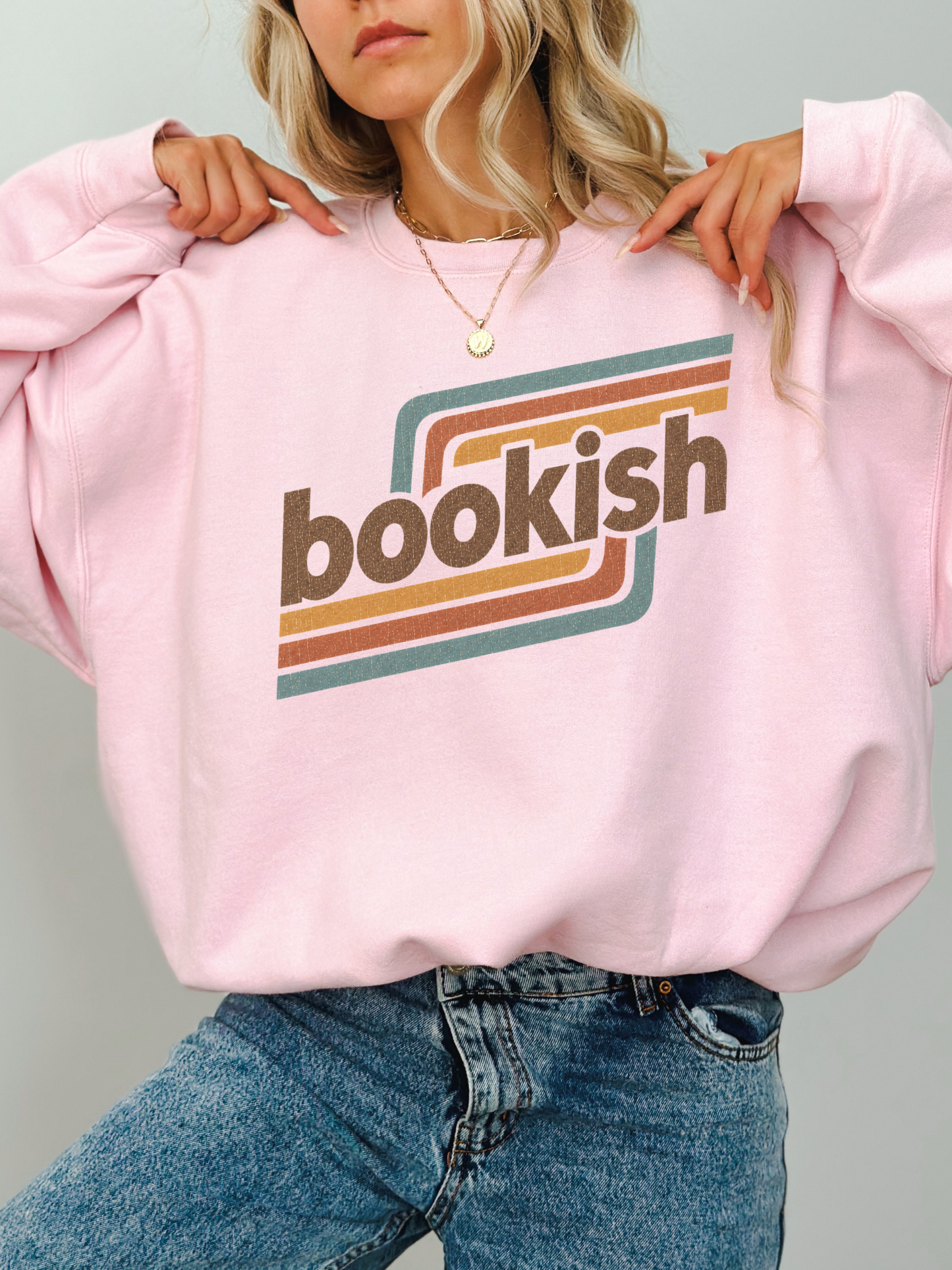 A woman wearing a light pink crewneck sweatshirt with a retro-style graphic that reads 'bookish' in brown lettering over teal and orange stripes, paired with blue jeans.