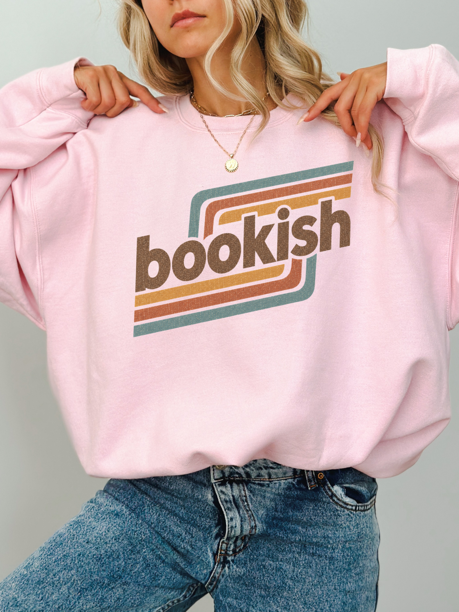 A woman wearing a light pink crewneck sweatshirt with a retro-style graphic that reads 'bookish' in brown lettering over teal and orange stripes, paired with blue jeans.