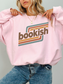 A woman wearing a light pink crewneck sweatshirt with a retro-style graphic that reads 'bookish' in brown lettering over teal and orange stripes, paired with blue jeans.