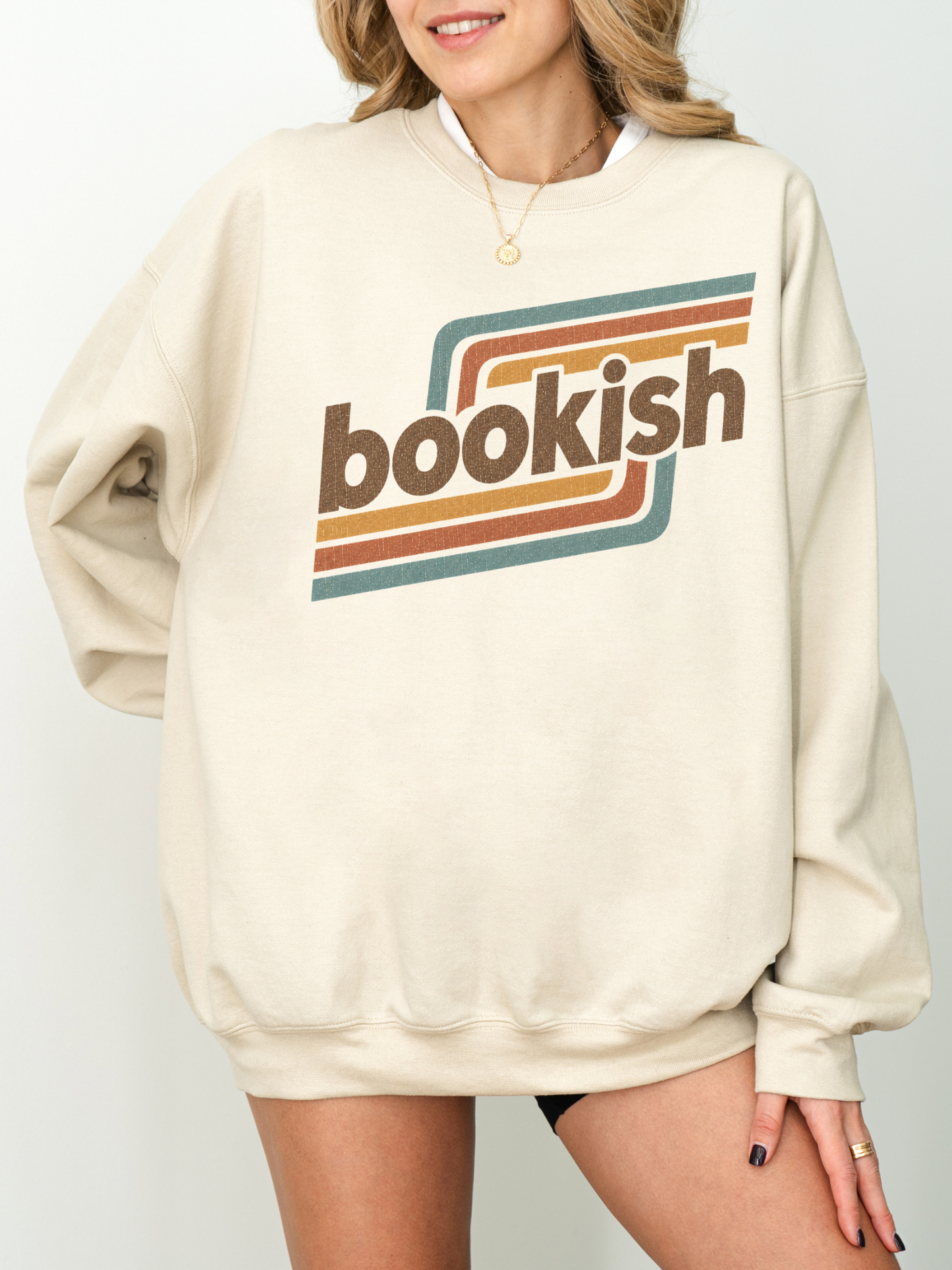 A woman wearing an oversized sand sweatshirt with a retro-style graphic on the chest that reads 'bookish' in brown lettering with orange, yellow, and teal stripes, set against a plain light gray background.