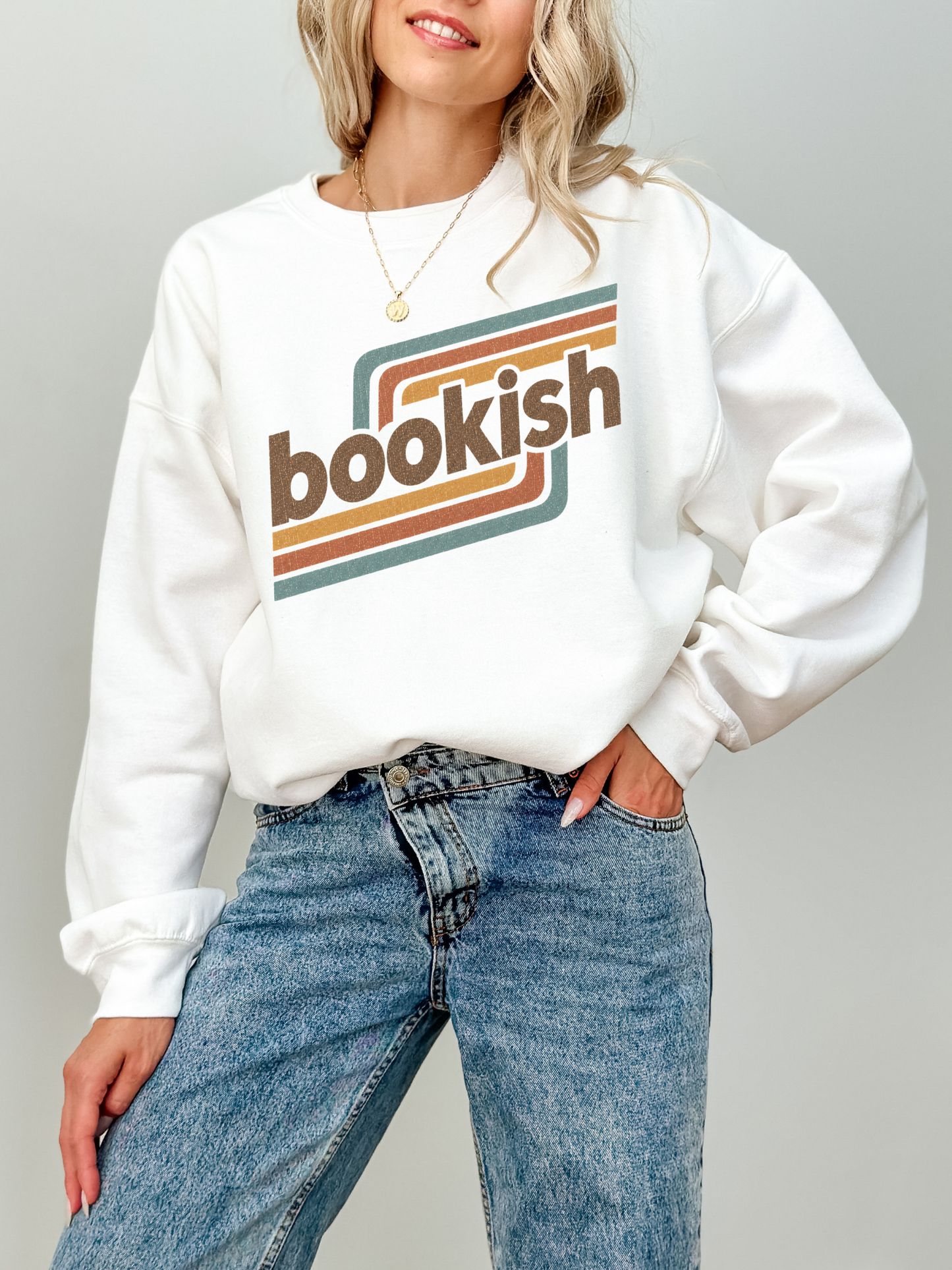 A woman wearing a white crewneck sweatshirt with a retro-style graphic that reads 'bookish' in brown text over orange and teal stripes, paired with blue jeans, against a light gray background.