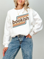 A woman wearing a white crewneck sweatshirt with a retro-style graphic that reads 'bookish' in brown text over orange and teal stripes, paired with blue jeans, against a light gray background.