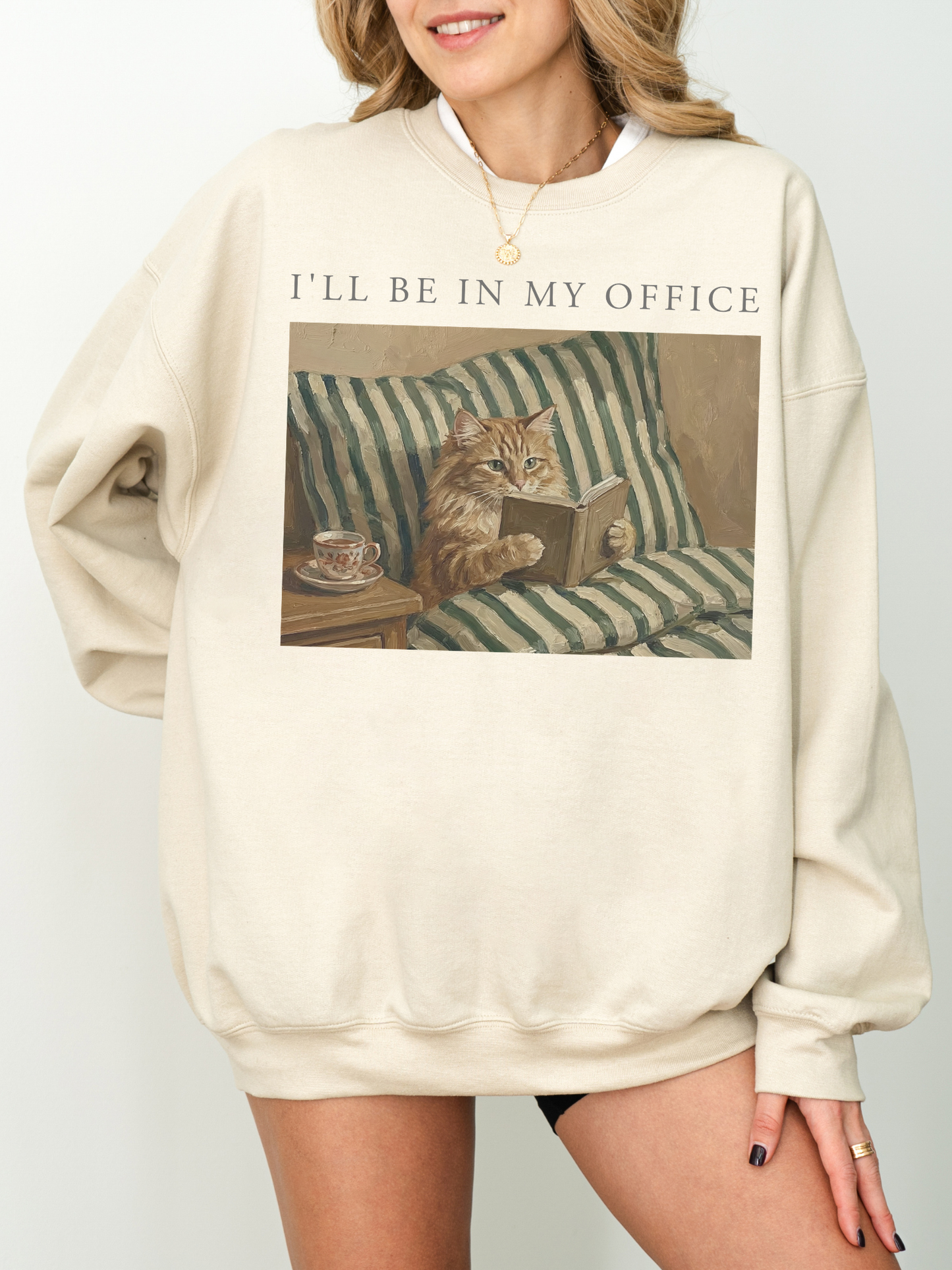 Sand oversized sweatshirt with Ill be in my office text and a cozy cat reading a book illustration with striped bedding and tea cup detail. Neutral aesthetic outfit for book lovers, introverts, and cozy reading days.