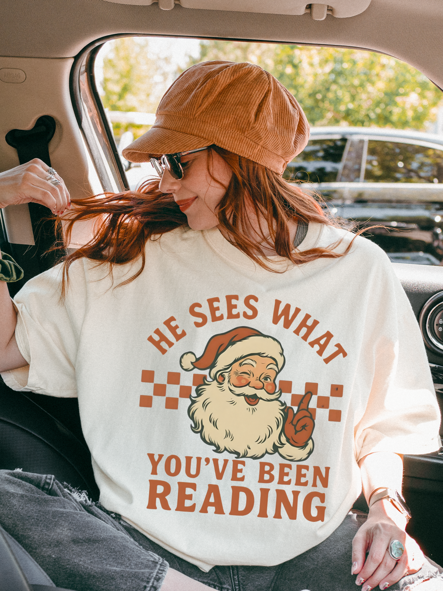 Woman in Ivory Santa reader shirt relaxing in car with sunglasses and brown cap, cozy bookish Christmas vibes