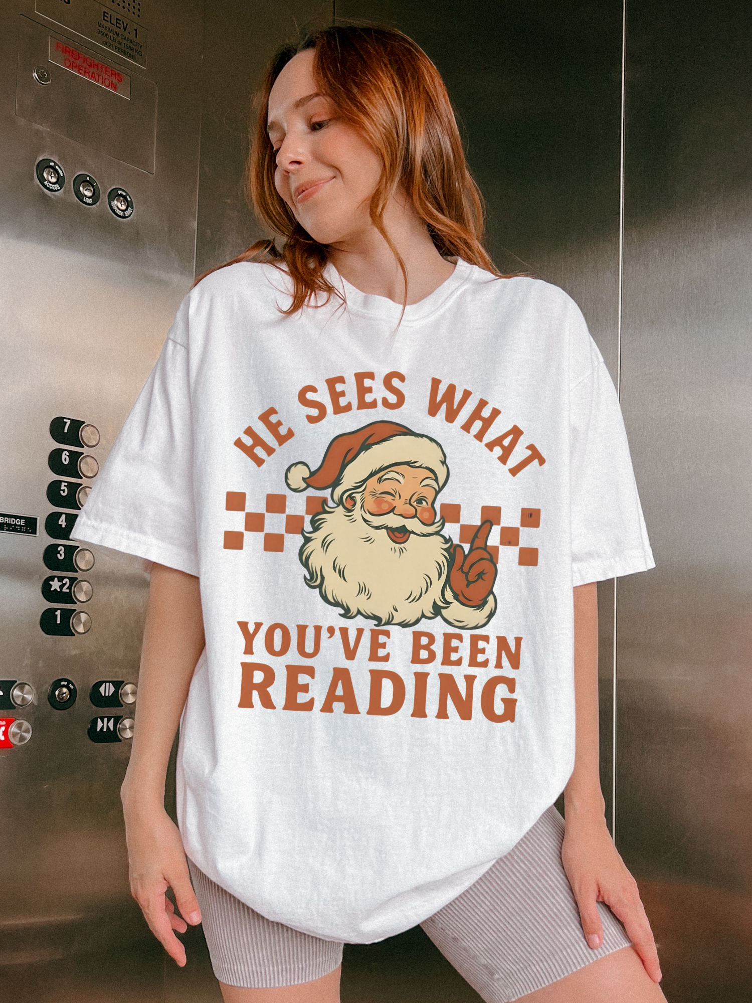 Woman in elevator wearing white Christmas shirt with cheeky Santa reading quote and biker shorts