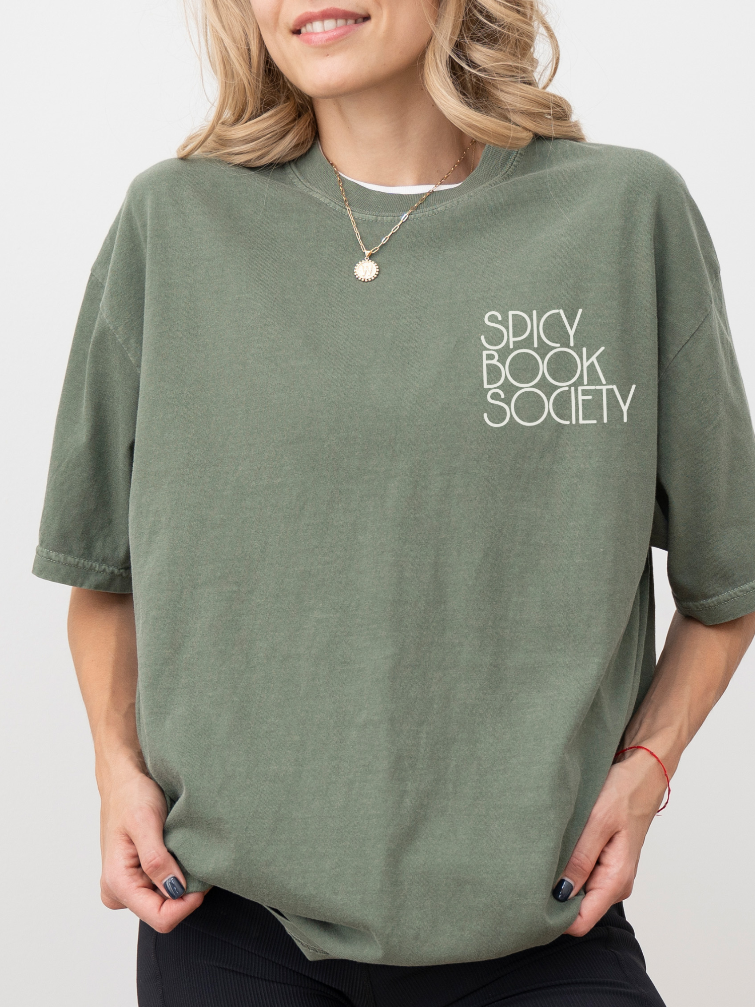 Woman wearing a moss green Comfort Colors T-shirt with "Spicy Book Society" printed on the left chest. She’s styled in casual bookish fashion with gold jewelry and soft waves, showcasing a cozy tee perfect for readers who love spicy books.