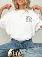 Woman sitting cross-legged wearing a white Comfort Colors T-shirt that says “Spicy Book Society.” Styled with jeans and layered gold necklaces in a cozy, bookish setting perfect for romantasy and smut readers.