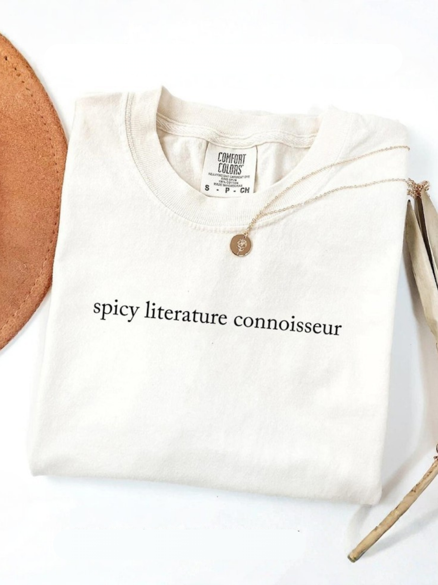 folded ivory Comfort Colors shirt with spicy literature connoisseur text and gold necklace flat lay