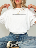 Woman in white Comfort Colors shirt sitting cross-legged in jeans and sneakers, shirt reads “spicy literature connoisseur.”