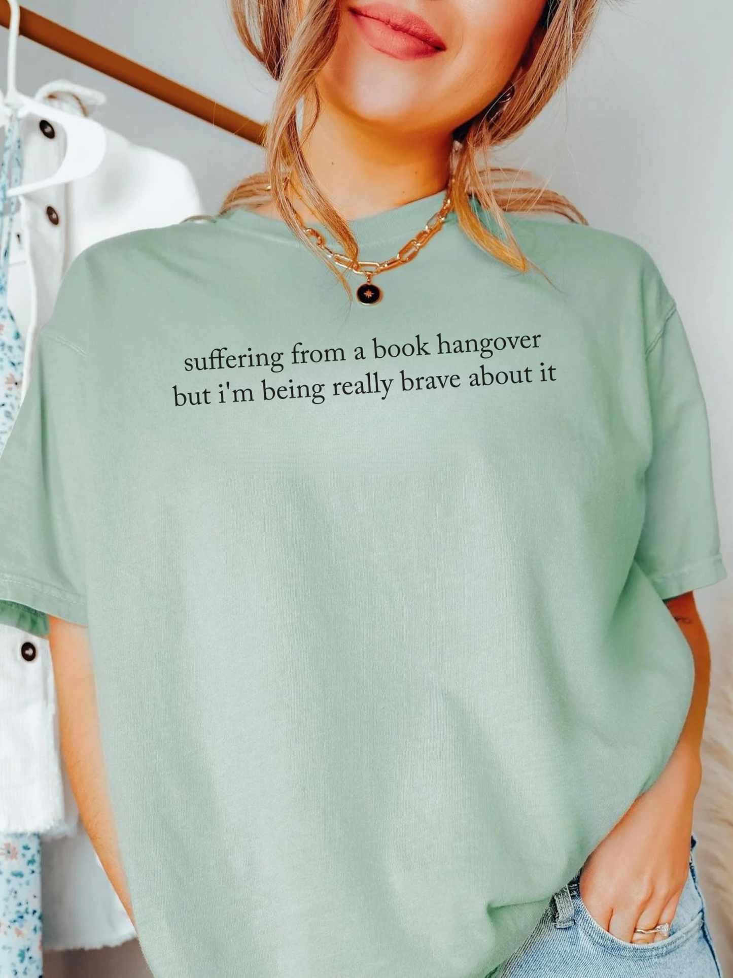 Woman wearing bay green Comfort Colors shirt that reads suffering from a book hangover but i'm being really brave about it styled with gold necklace and casual outfit in bright indoor setting