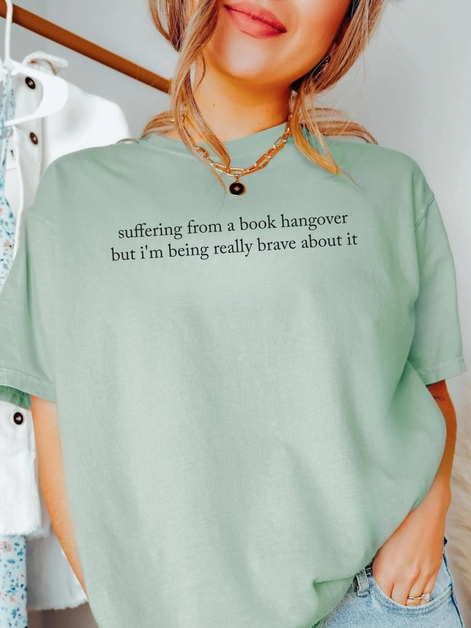 Woman wearing bay green Comfort Colors shirt that reads suffering from a book hangover but i'm being really brave about it styled with gold necklace and casual outfit in bright indoor setting