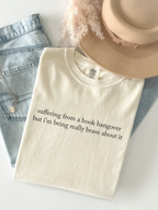 Ivory Comfort Colors book hangover shirt folded in flatlay with jeans beige hat and jewelry featuring minimalist text suffering from a book hangover but i'm being really brave about it
