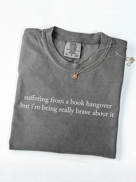 Pepper Comfort Colors book lover shirt folded with necklace in flatlay showing minimalist quote suffering from a book hangover but i'm being really brave about it
