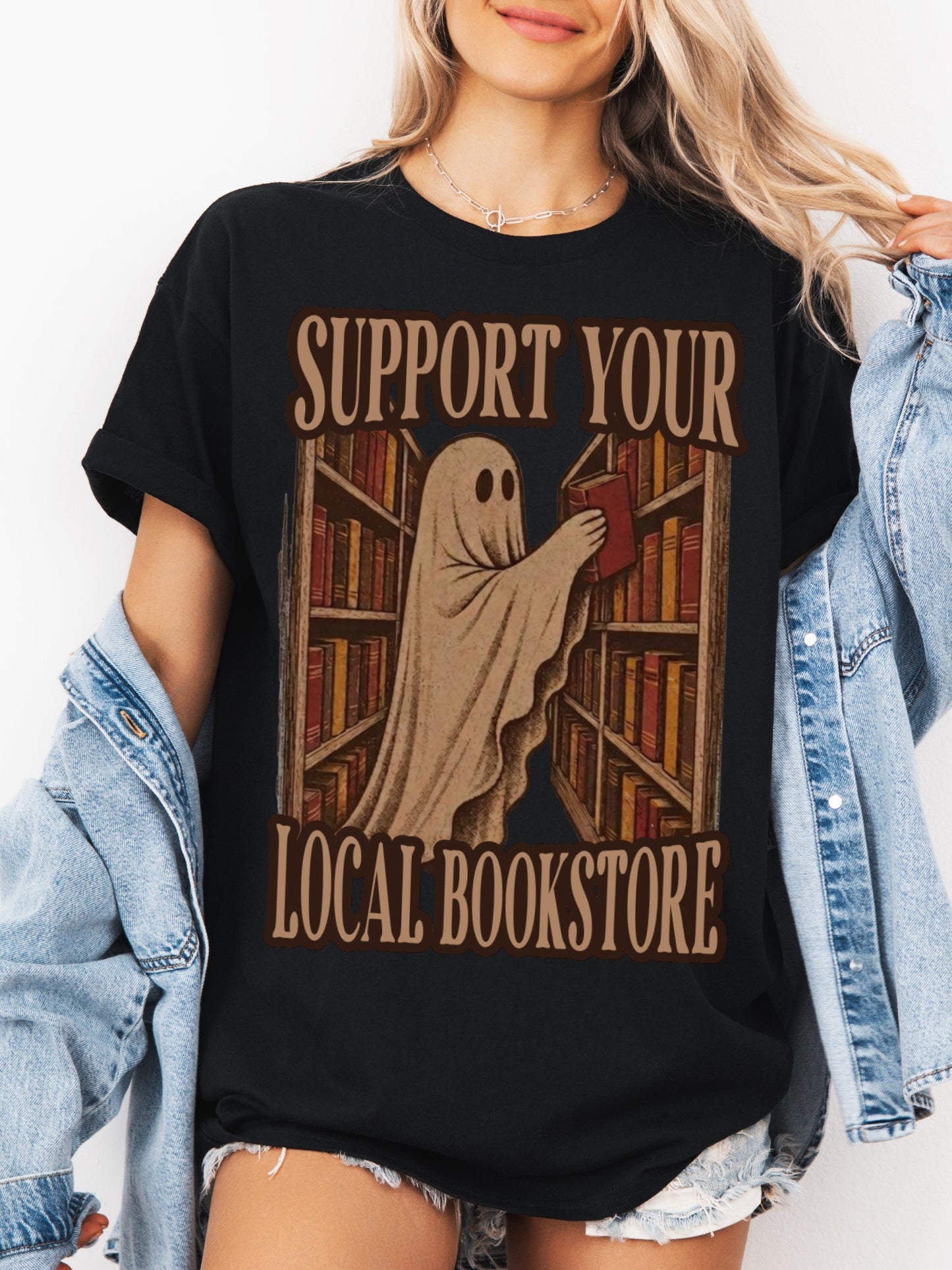A woman wearing a black graphic t-shirt with a 'Support Your Local Bookstore' ghost and bookshelf design, styled with an open light blue denim jacket.