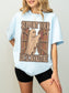 A woman wearing an oversized chambray graphic T-shirt with a ghost reading books design and the text 'SUPPORT YOUR LOCAL BOOKSTORE', paired with black shorts against a light gray studio background.