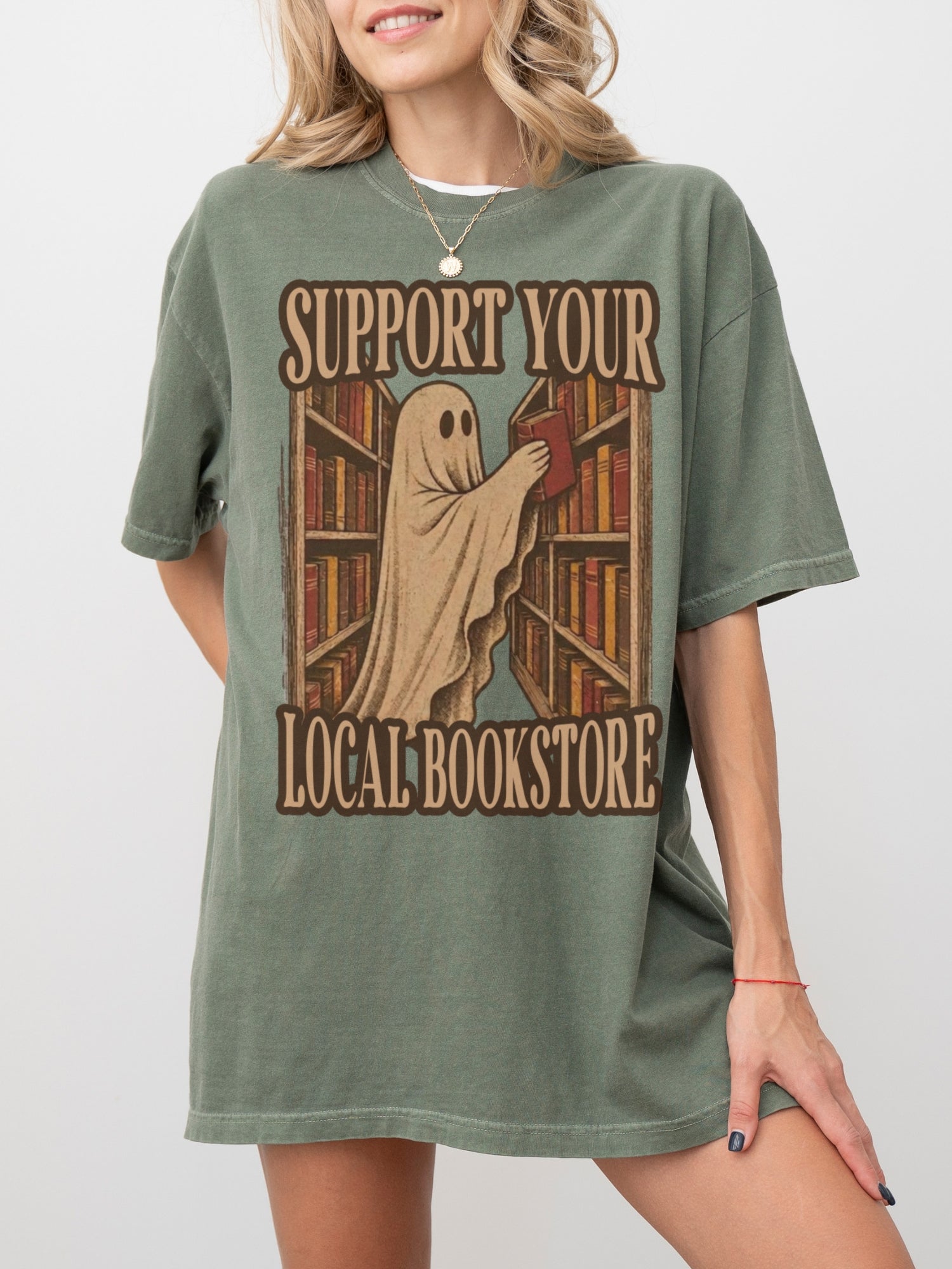 A woman wearing an oversized moss green graphic t-shirt with a vintage-style print of a ghost reading a book in a library, with the text 'SUPPORT YOUR LOCAL BOOKSTORE'.