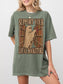 A woman wearing an oversized moss green graphic t-shirt with a vintage-style print of a ghost reading a book in a library, with the text 'SUPPORT YOUR LOCAL BOOKSTORE'.