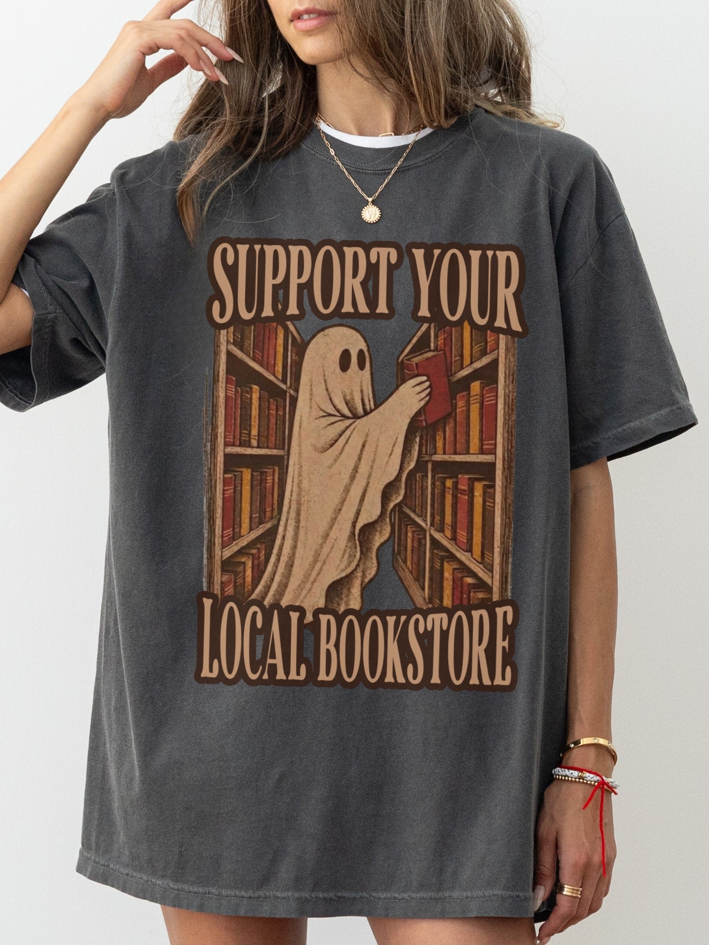 A woman wearing an oversized pepper T-shirt with a ghost reading books at a library and the text 'SUPPORT YOUR LOCAL BOOKSTORE'.