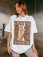 A woman wearing an oversized white graphic t-shirt with a 'Support Your Local Bookstore' ghost and bookshelf design, standing inside an elevator.