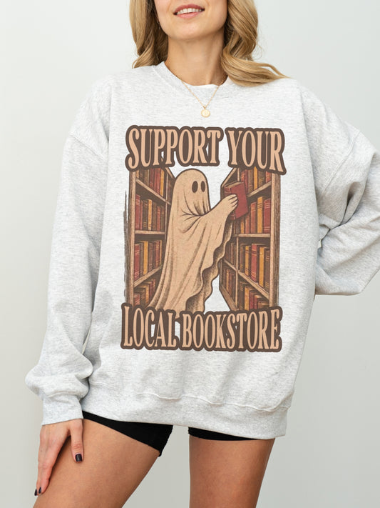 A woman wearing an ash heather gray sweatshirt with a graphic print of a ghost reading books at a bookshelf, with the text 'SUPPORT YOUR LOCAL BOOKSTORE'.
