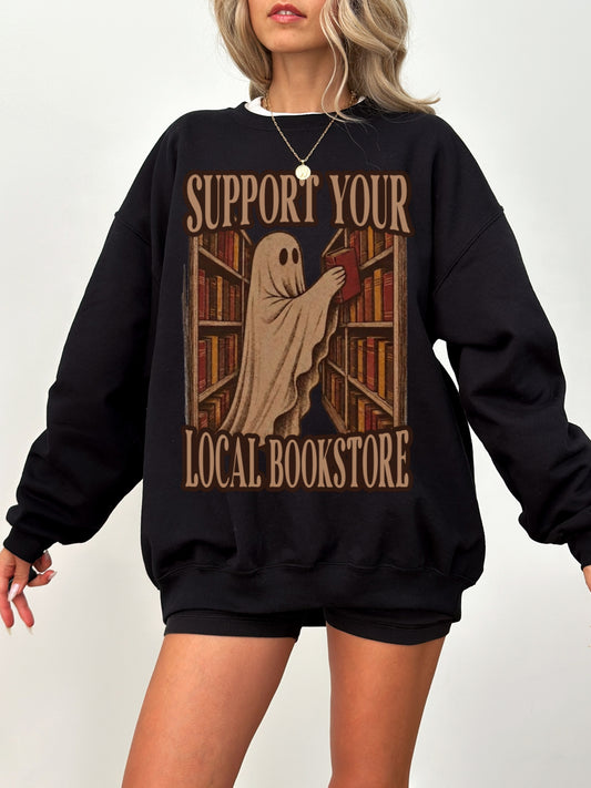 A woman wearing a black crewneck sweatshirt with a graphic print that reads 'SUPPORT YOUR LOCAL BOOKSTORE' featuring a ghost holding a book in front of bookshelves.