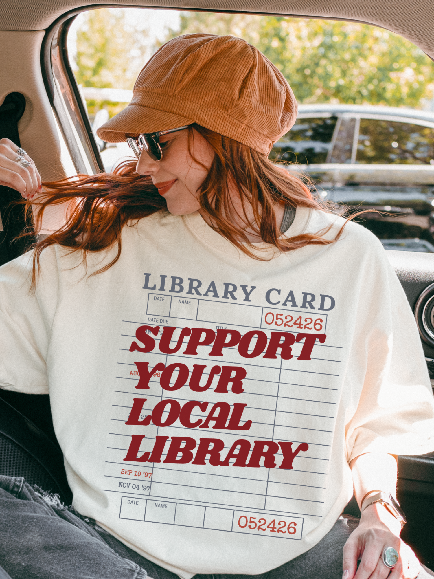 woman sitting in car wearing cream support your local library t shirt with vintage library card design and casual fall outfit