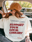 woman sitting in car wearing cream support your local library t shirt with vintage library card design and casual fall outfit