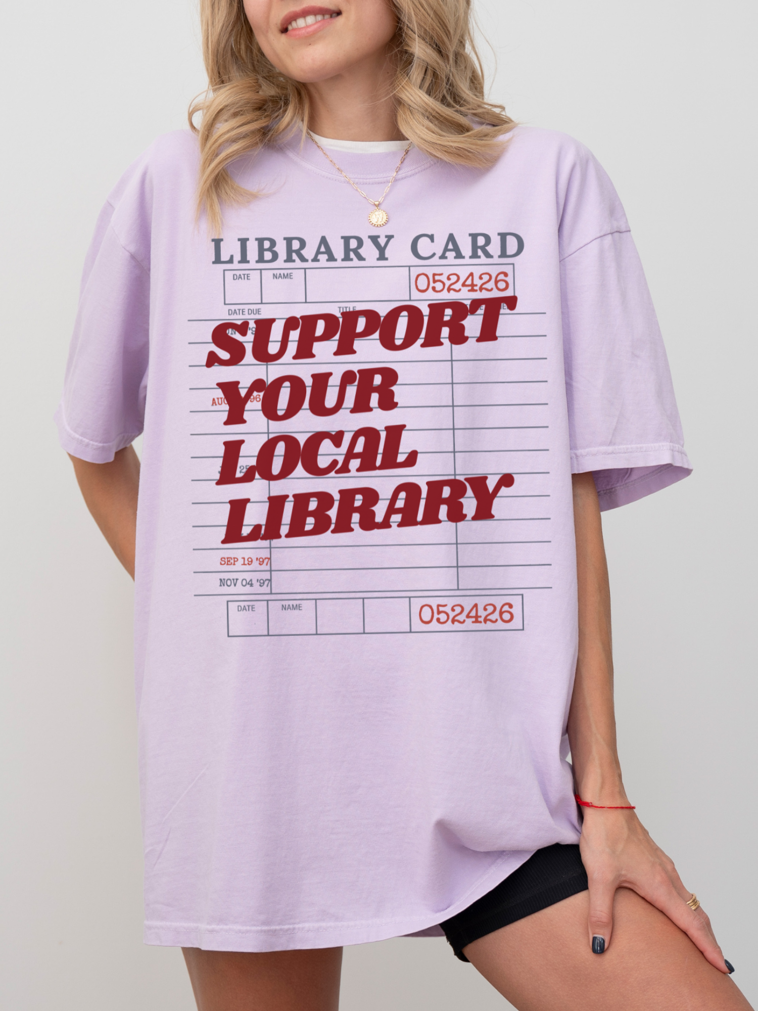 woman wearing oversized orchid t shirt with support your local library text and retro library card design for cozy book lover aesthetic