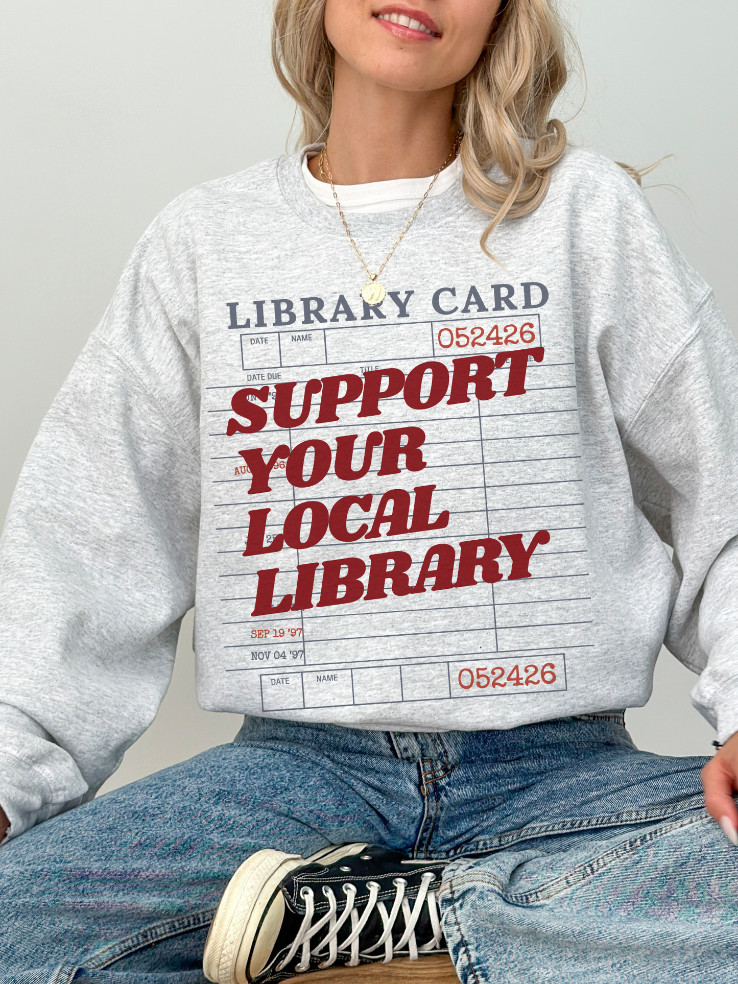 woman wearing ash grey sweatshirt with support your local library text and retro library card graphic styled with jeans and sneakers casual look