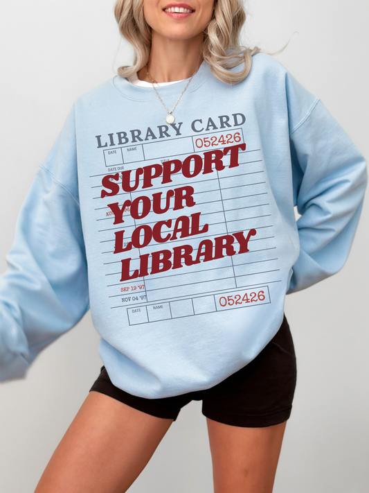 woman wearing light blue sweatshirt with support your local library text and vintage library card graphic styled casually for book lovers