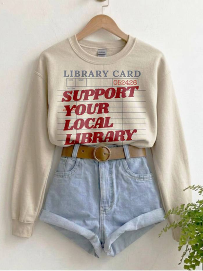cream sweatshirt featuring support your local library text with vintage library card design styled with denim shorts and neutral tones
