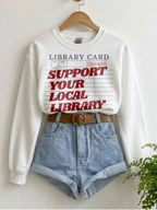 white sweatshirt with support your local library graphic styled with denim shorts and belt on hanger in minimal aesthetic setting