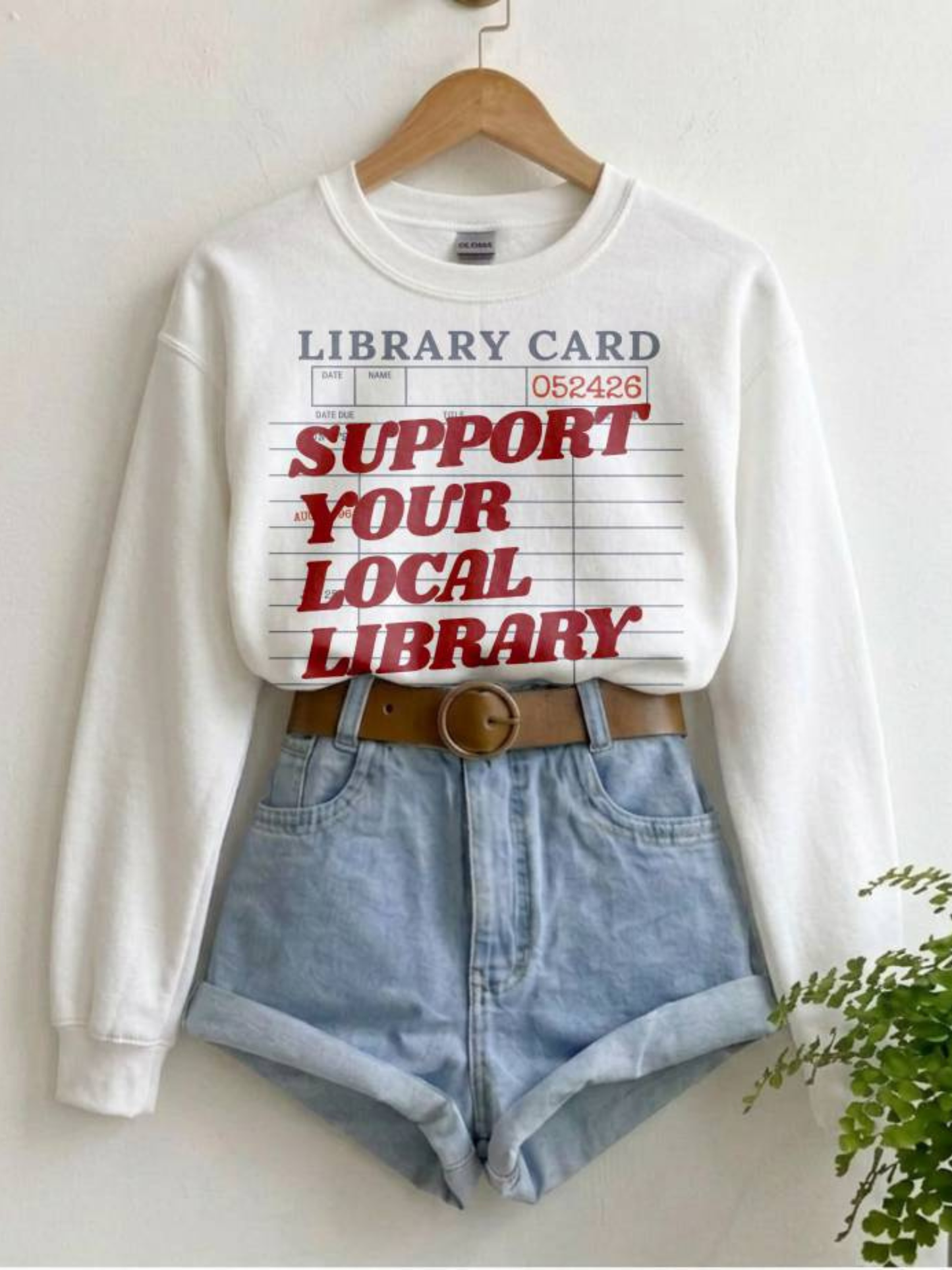 white sweatshirt with support your local library graphic styled with denim shorts and belt on hanger in minimal aesthetic setting