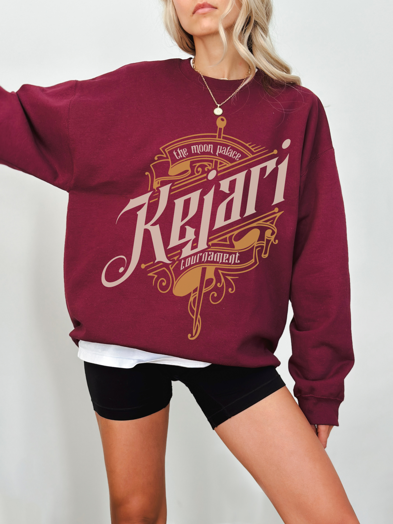 Maroon sweatshirt featuring ornate gold and tan typography reading Kejari Tournament, modeled with black biker shorts and casual layered top, inspired by fantasy book fandom.