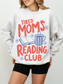 woman wearing ash grey tired moms reading club sweatshirt with colorful retro typography coffee mug and open book design for readers