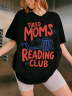woman wearing oversized black tired moms reading club graphic t shirt with coffee mug open book and floral reading design in elevator