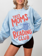 woman wearing oversized light blue tired moms reading club sweatshirt with retro typography coffee mug floral accents and open book artwork