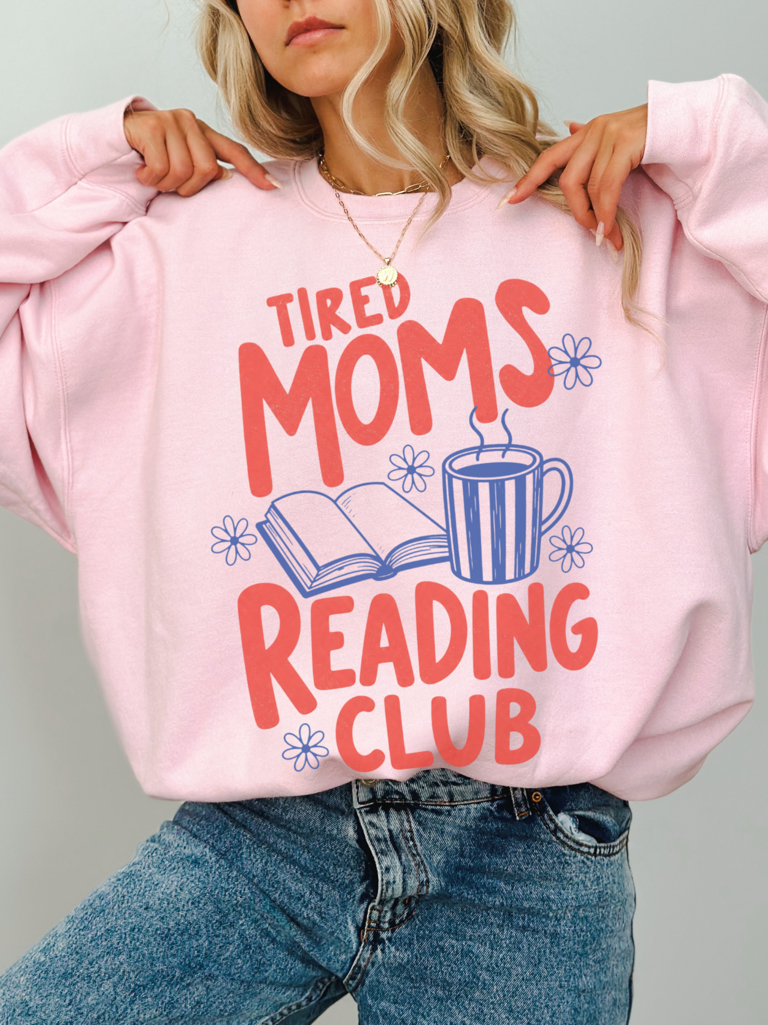 woman wearing oversized light pink tired moms reading club sweatshirt with colorful reading graphic coffee mug and open book illustration
