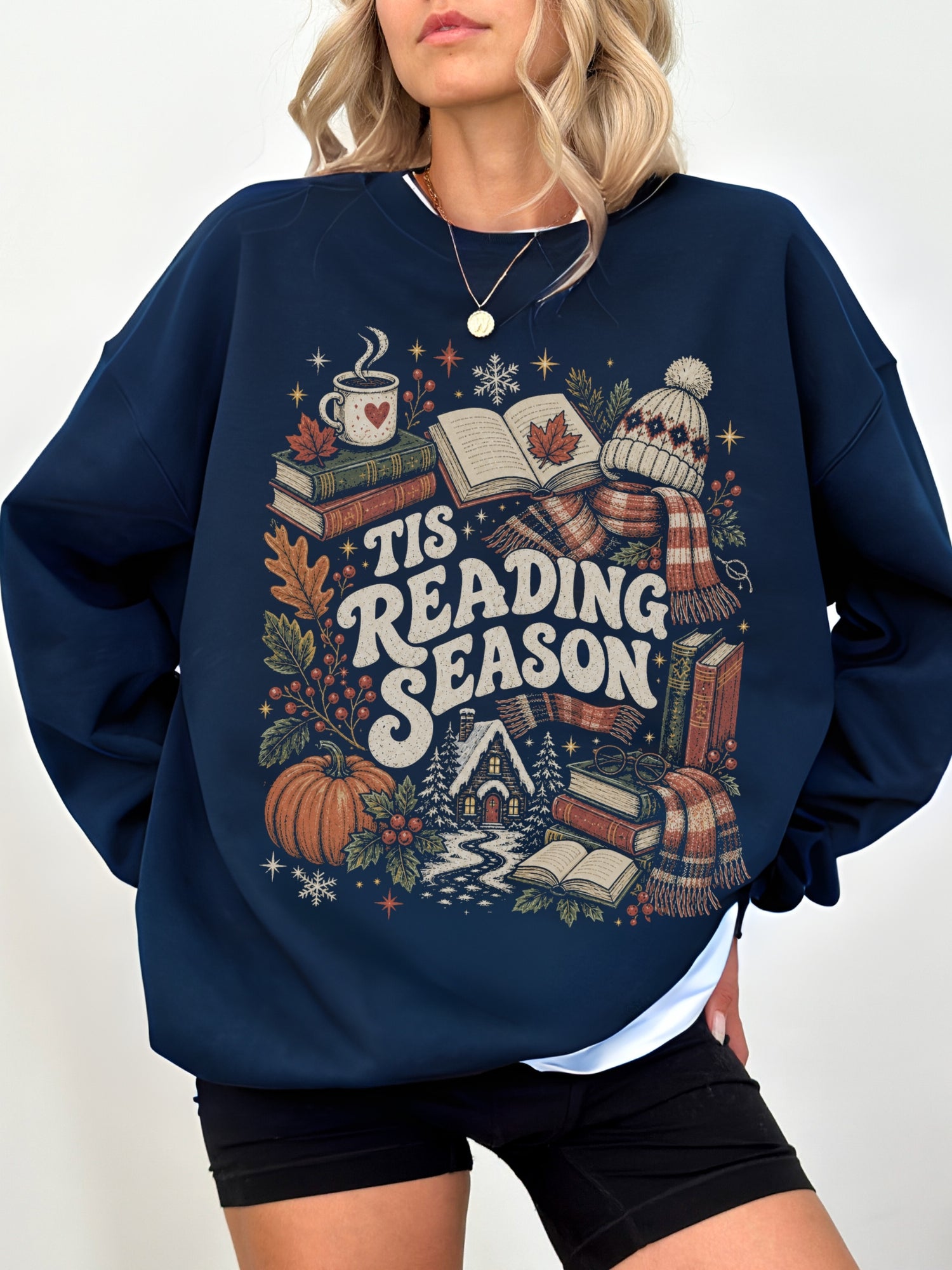 Navy Gildan sweatshirt modeled with a Tis Reading Season design featuring stacked books, open book, coffee mug, autumn leaves, pumpkin, scarf and snowy cottage, cozy reading sweatshirt for book lovers