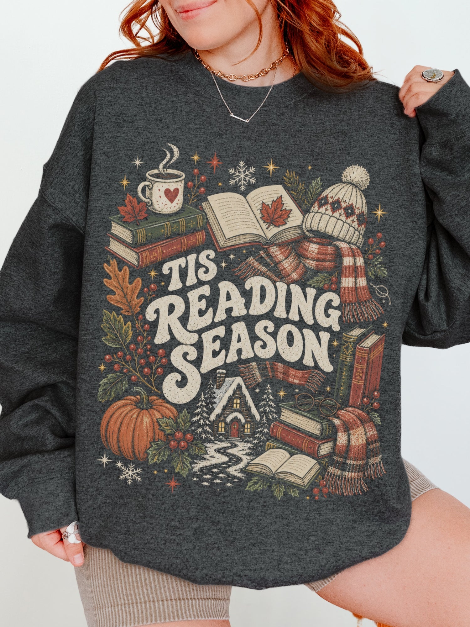 Dark Grey Heather Gildan sweatshirt featuring a cozy Tis Reading Season graphic with books, coffee, pumpkin, fall foliage, winter scarf, knit hat and snowy cottage, seasonal bookish sweatshirt for readers