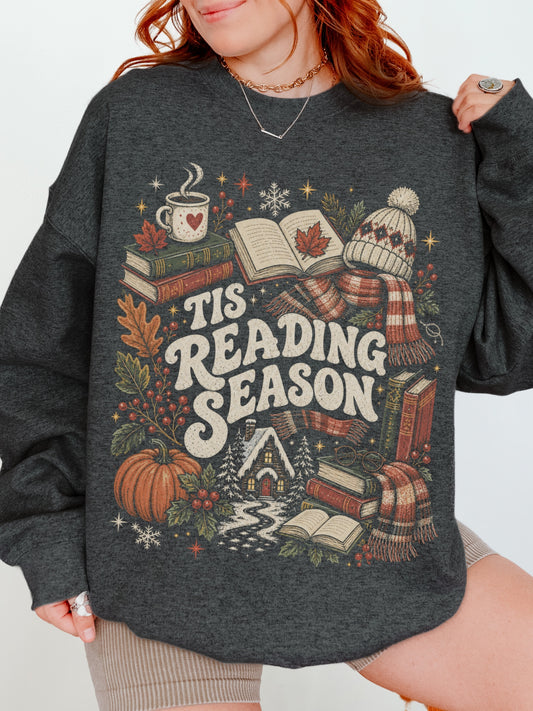 Dark Grey Heather Gildan sweatshirt featuring a cozy Tis Reading Season graphic with books, coffee, pumpkin, fall foliage, winter scarf, knit hat and snowy cottage, seasonal bookish sweatshirt for readers